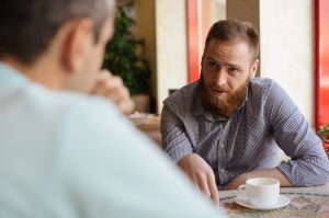 photo of two men chatting around a table. One man's face can be scene, other man's back is in image