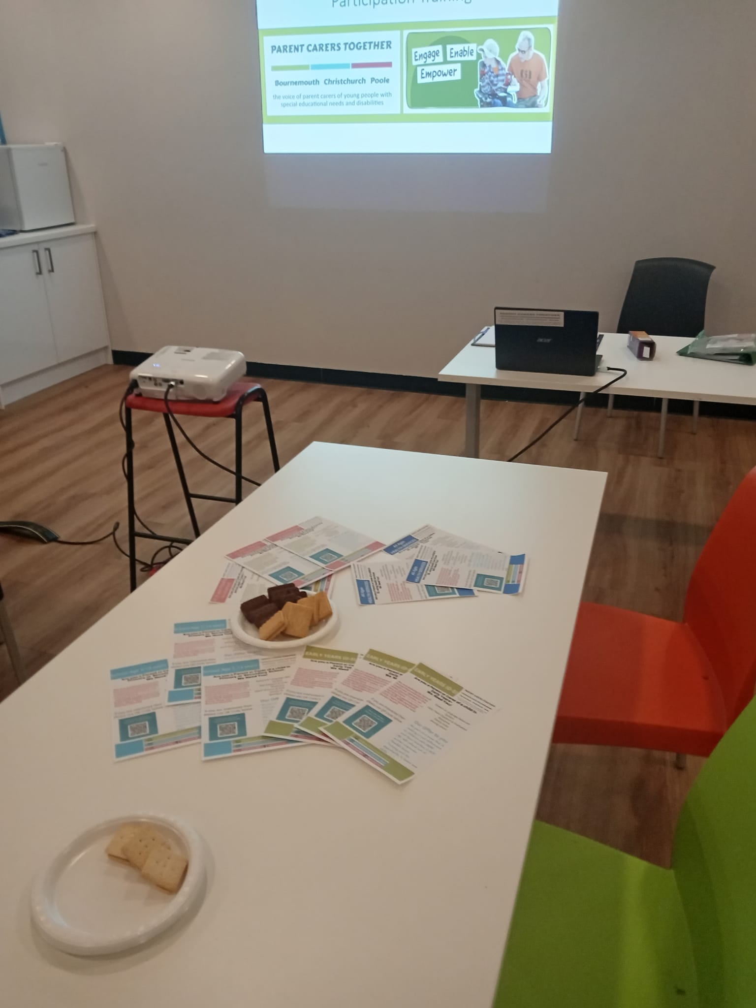room set up for training-including tables, chairs, a projector, leaflets, biscuits and screen on a wall.